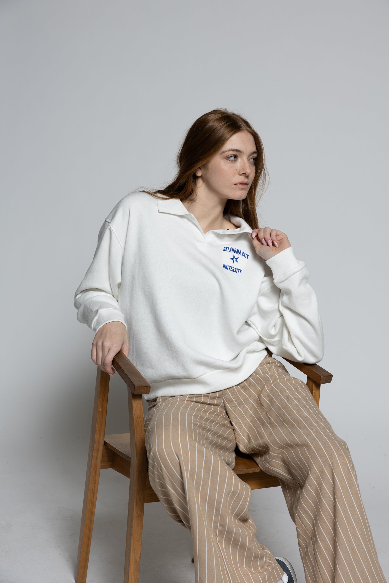 Woman sitting on a chair wearing a white sweatshirt with a logo and beige pants against a gray background