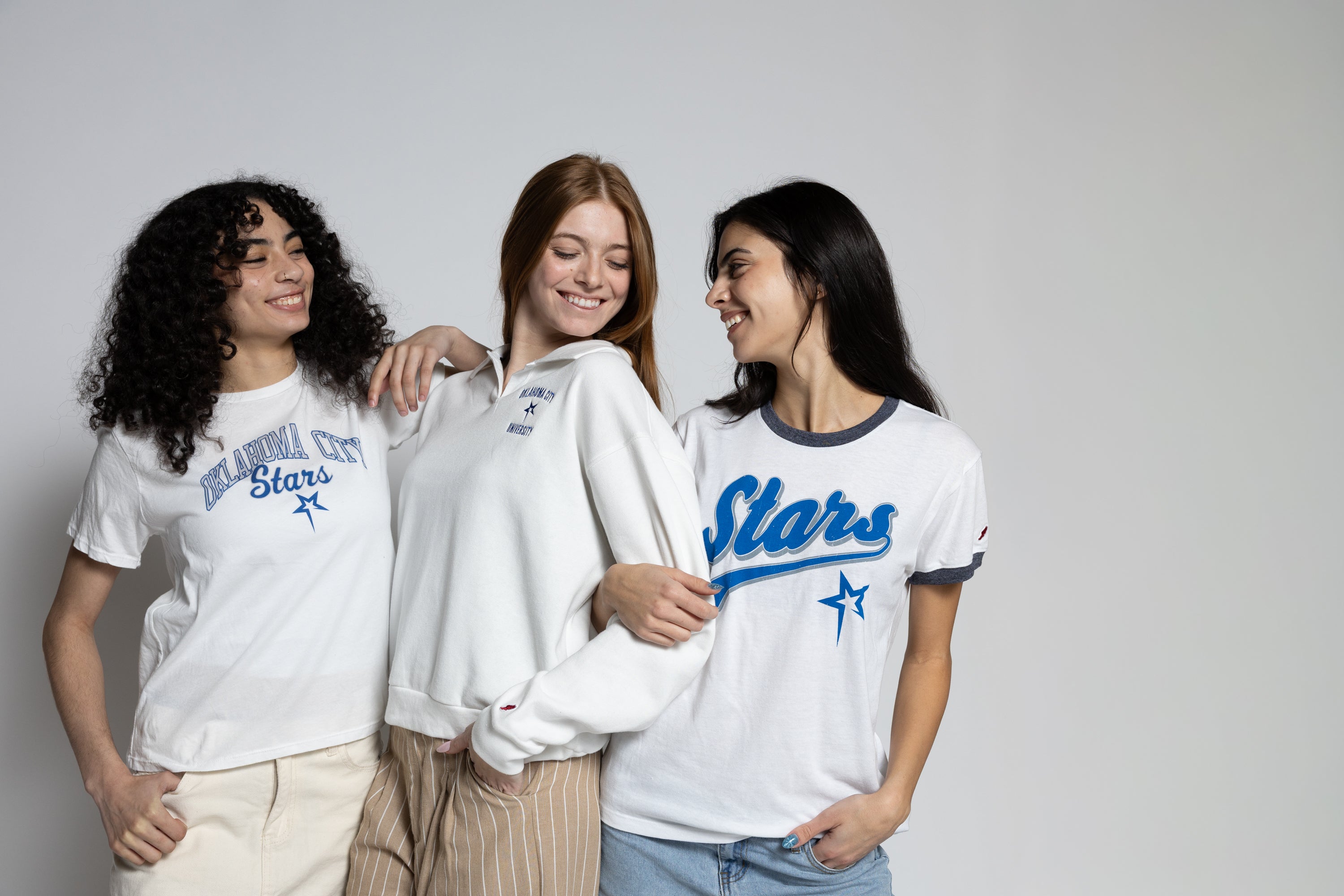 Three women wearing OCU Stars t-shirts and sweatshirts on a gray background